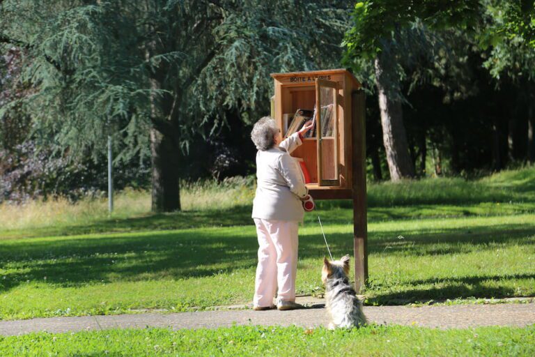 Une dame accompagnée de son chien en train de chercher dans une boîte à livres